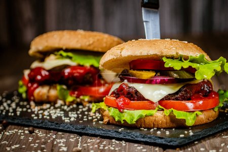 Großaufnahme von leckeren Burgern mit Rindfleisch, Käse, Tomaten, Zwiebeln, Paprika und Salat auf dem Steinbrett. Selektiver Fokus. Burger mit Messer 