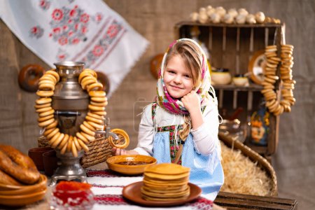 Foto de Cute little girl in a folk Russian headscarf and national dress with bunch of bagels and pancakes celebrating Maslenisa. Traditional Russian food for Shrovedite. - Imagen libre de derechos