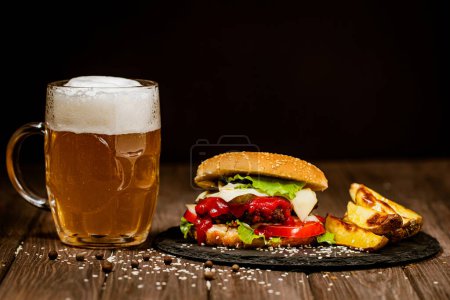 Photo for Side view of homemade beef burgers with mug of beer, baked potato tomatoes, cheese, sauce on black background. Selective focus. Delicious food. - Royalty Free Image