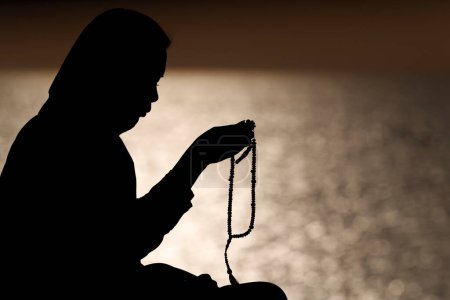 Téléchargez les photos : Silhouette de femme musulmane tenant des perles de prière dans ses mains et priant au coucher du soleil. Religion concept de prière - en image libre de droit
