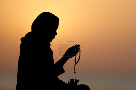 Téléchargez les photos : Silhouette de femme musulmane tenant des perles de prière dans ses mains et priant au coucher du soleil. Religion concept de prière - en image libre de droit