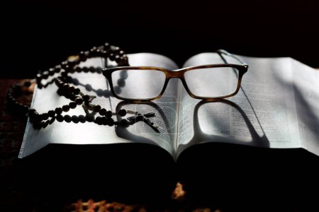 Foto de Biblia abierta con un par de gafas y un rosario. Candeles de iglesia en la iglesia. Mujer rezando a la Virgen. - Imagen libre de derechos