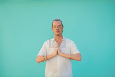 Photo for Tattooed male in white shirt and bracelet practicing yoga with closed eyes and namaste hands - Royalty Free Image