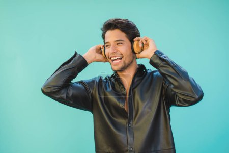 Photo for Happy male in black shirt listening to music from headphones while looking away on aquamarine background - Royalty Free Image