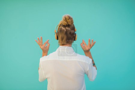 Foto de Vista posterior de mujer tatuada anónima en auriculares inalámbricos escuchando la canción mientras muestra mudra de sabiduría sobre fondo aguamarina - Imagen libre de derechos
