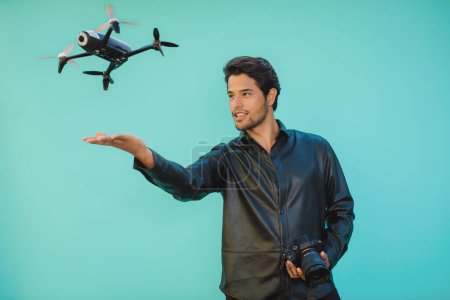 Téléchargez les photos : Young unshaven male in black apparel with professional photo camera and flying UAV representing bird concept on aquamarine background - en image libre de droit