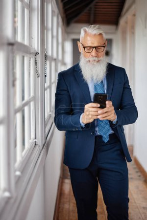 Téléchargez les photos : Exécutif masculin bien habillé avec message texte barbe grise sur téléphone portable tout en restant debout dans le passage - en image libre de droit