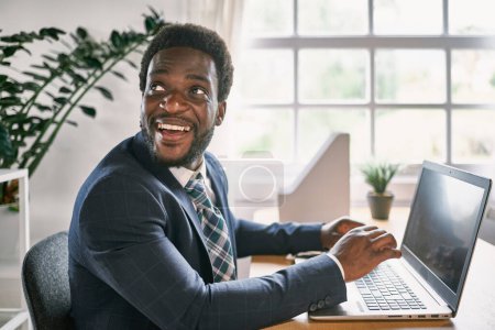 Téléchargez les photos : Smiling African American male employee looking away at desk with netbook in workspace on sunny day - en image libre de droit