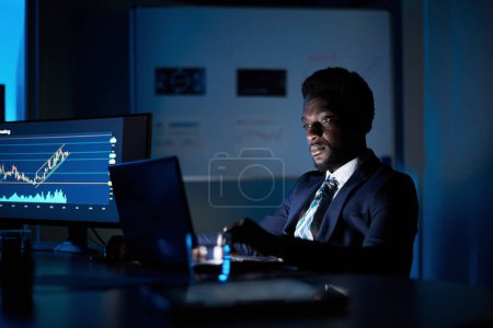 Photo for Serious African American male analyst in formal wear working on netbook against graphics on monitor in workspace - Royalty Free Image