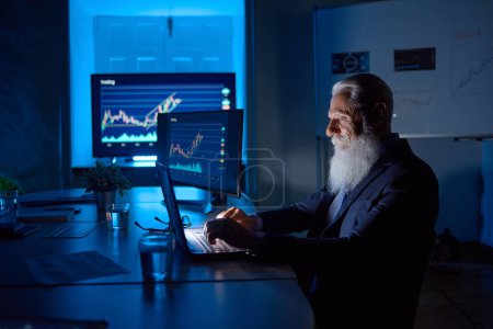 Téléchargez les photos : Vue latérale de l'analyste masculin âgé avec barbe grise travaillant sur netbook à la table avec des graphiques sur moniteurs dans l'espace de travail - en image libre de droit