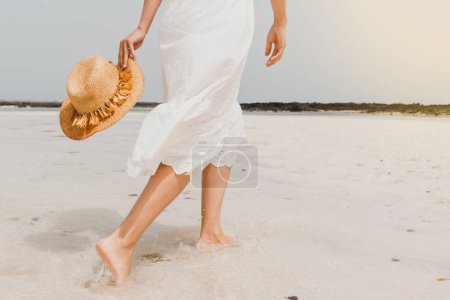 Foto de Vista posterior de la cosecha anónima hembra descalza en vestido blanco y con sombrero de paja en la mano paseando en la playa de arena húmeda en el día de verano - Imagen libre de derechos
