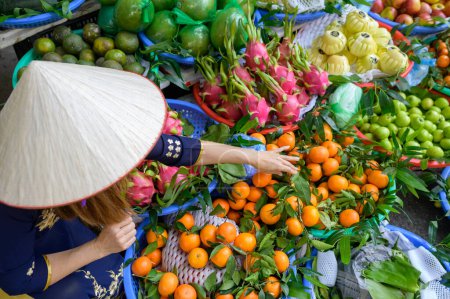 Téléchargez les photos : D'en haut de femelle méconnaissable en vêtements traditionnels et chapeau conique asiatique debout dans le marché alimentaire local avec assortiment de fruits exotiques colorés au Vietnam - en image libre de droit