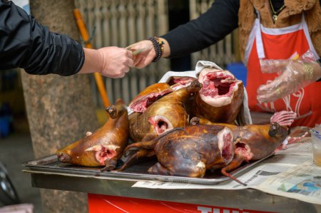 Téléchargez les photos : Des carcasses de chiens rôtis placées sur la table devant un acheteur anonyme payant au vendeur de cultures sur le marché vietnamien local - en image libre de droit