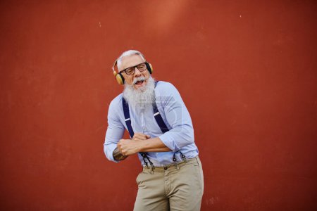 Foto de Hombre hipster senior en baile de moda mientras escucha la canción de los auriculares y mira hacia otro lado en el fondo rojo - Imagen libre de derechos