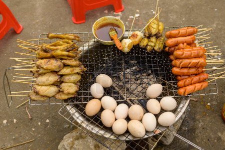 Téléchargez les photos : Grill en métal avec des ailes de poulet rôties et les pieds placés près des saucisses et des pommes de terre sur des bâtons contre les œufs et bol de sauce sur brasero sur la rue Vietnam - en image libre de droit