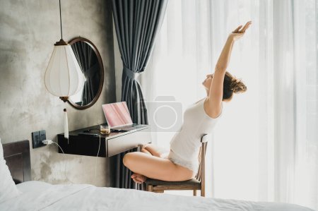 Téléchargez les photos : Vue latérale complète du corps de la jeune femme en vêtements de nuit assis avec les jambes croisées sur la chaise à la table avec tasse de boisson et ordinateur tout en s'étirant le matin - en image libre de droit