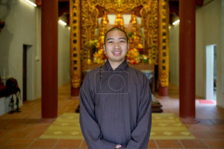 Foto de Sonriente monje asiático vestido de negro mirando a la cámara mientras está de pie dentro del templo con estatua de oro en Vietnam - Imagen libre de derechos