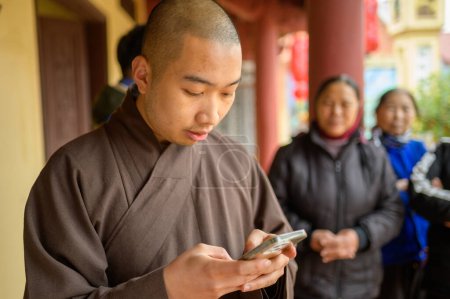 Téléchargez les photos : Jeune homme asiatique en robe noire du monastère debout sur la rue près du temple et en utilisant smartphone - en image libre de droit