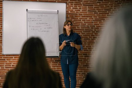 Photo for Serious adult businesswoman in eyeglasses standing near whiteboard and talking during seminar in spacious room with brick wall - Royalty Free Image