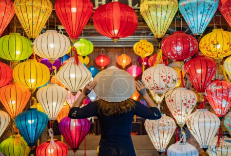 Téléchargez les photos : Vue de dos femme anonyme en robe touchant chapeau conique asiatique sur la tête tout en se tenant près de lanternes colorées suspendues au plafond - en image libre de droit