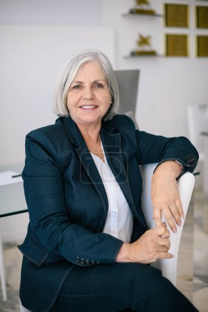 Photo for Smiling mature female entrepreneur with gray hair in formal suit sitting on chair and looking at camera in light room - Royalty Free Image