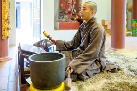Téléchargez les photos : Jeune homme asiatique en robe noire assis dans le temple et jouer cloches bol pendant le service - en image libre de droit