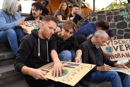 Téléchargez les photos : Groupe de manifestants concentrés et diversifiés créant des pancartes en carton assis sur les escaliers pendant la préparation de la grève - en image libre de droit