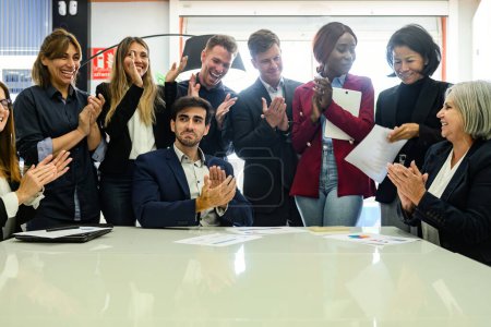 Photo for Group of cheerful diverse colleagues in formal clothes standing at table in office and applauding while celebrating success - Royalty Free Image