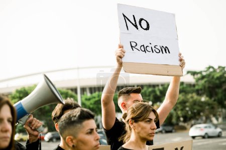 Téléchargez les photos : D'en bas un groupe de jeunes gens en vêtements décontractés debout avec le bras levé et tenant un signe avec l'inscription Pas de racisme. - en image libre de droit