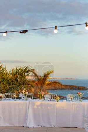 Téléchargez les photos : Paysage pittoresque de tables de banquet avec des fleurs et des chaises contre les palmiers exotiques et la mer sous le ciel nuageux coucher de soleil et guirlande dans la station tropicale - en image libre de droit