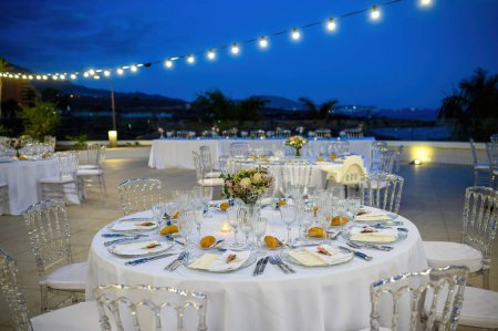 Téléchargez les photos : Tables rondes servies avec verrerie et fleurs sur nappe blanche près de chaises dans un restaurant contemporain décoré en plein air pendant la célébration du soir - en image libre de droit