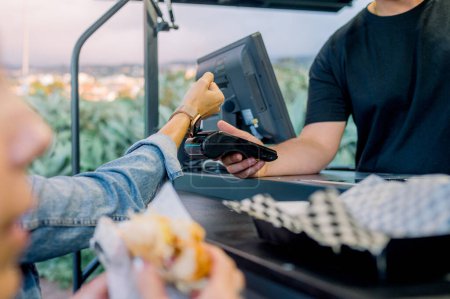 Téléchargez les photos : Femme de culture floue manger hot dog près du comptoir tandis que le client méconnaissable payer pour l'achat avec montre intelligente dans un café extérieur - en image libre de droit