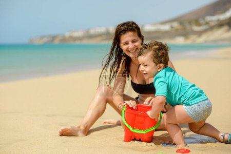 Téléchargez les photos : Plein corps vue latérale de heureux petit garçon et fille en vêtements décontractés souriant et regardant la caméra tout en jouant ensemble sur la plage de sable - en image libre de droit