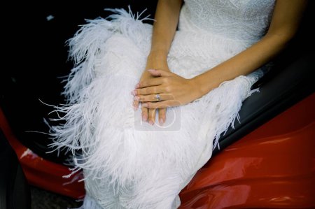 Hochwinkel der anonymen Braut im eleganten weißen Hochzeitskleid mit Federn und Ring sitzt während der Feier im modernen roten Auto