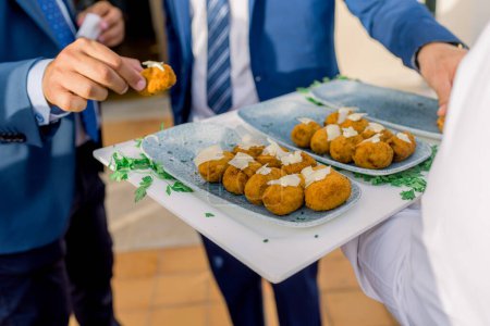 Anonymous male business partners in elegant suits taking appetizing potato croquettes from plate served with fresh herbs during banquet process in restaurant