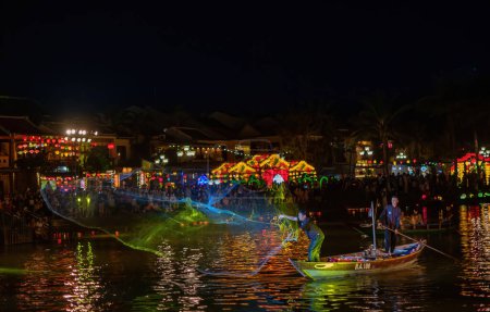 Foto de Pescadores irreconocibles con red de lanzamiento de barcos sobre el agua por la noche en la orilla de la ciudad con multitud de personas con luces multicolores y globos celebrando el carnaval - Imagen libre de derechos