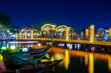 Photo for Amazing view of yellow and blue glowing lights on exterior of buildings and bridge reflective over calm river with canoes at night under blue sky - Royalty Free Image