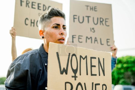Téléchargez les photos : Portrait de femme sérieuse en vêtements décontractés debout près de l'affiche tout en tenant les femmes placard de puissance et en regardant loin en plein air lumière du jour - en image libre de droit