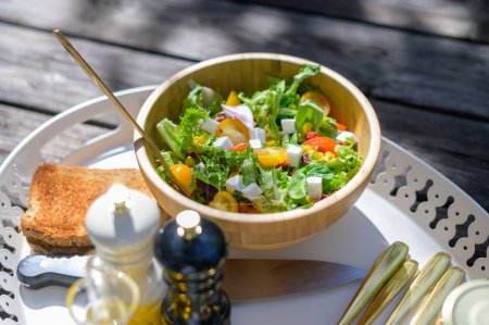 Photo for From above of appetizing healthy salad with greens cherry tomatoes and Feta cheese served in wooden bowl on gray with crispy toast and cutlery - Royalty Free Image