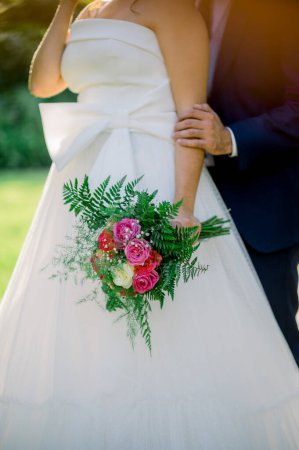 Photo for Faceless bride in white strapless dress with bow holding colorful bouquet standing with loving groom behind back in sunlight - Royalty Free Image