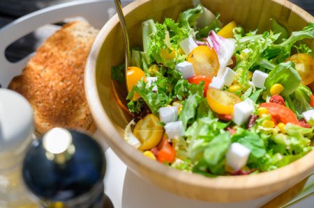 Téléchargez les photos : Haut angle de salade fraîche appétissante avec tomates cerises à la laitue et fromage Feta servi dans un bol en bois placé sur un plateau près de pain grillé croustillant - en image libre de droit