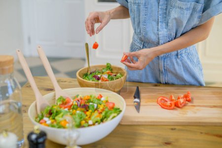Photo for Crop unrecognizable female in denim overall standing near counter and adding cut tomatoes into bowl with fresh healthy salad while preparing lunch in kitchen - Royalty Free Image