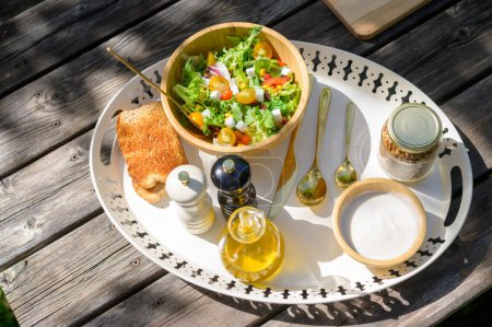 Foto de Vista superior del tazón con ensalada saludable servida en bandeja con tostadas crujientes y yogur cerca del frasco de granola y cubiertos en un día soleado - Imagen libre de derechos
