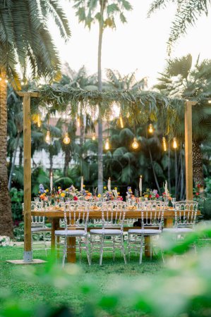 Photo for Wooden table with arch decorated with plants and hanging light bulbs and transparent chairs ready for wedding banquet in tropical resort - Royalty Free Image
