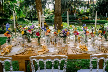 Foto de Conjunto de mesa de madera con exquisitos cubiertos y decorado con flores brillantes y velas en el jardín del exótico hotel ecológico - Imagen libre de derechos