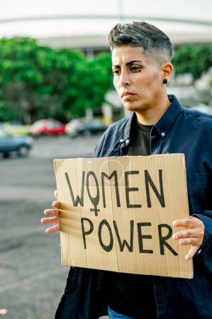 Foto de Mujer seria con el pelo corto mostrando la pancarta de poder de las mujeres y mirando hacia otro lado mientras está de pie en la calle y protestando por las mujeres en el parque - Imagen libre de derechos
