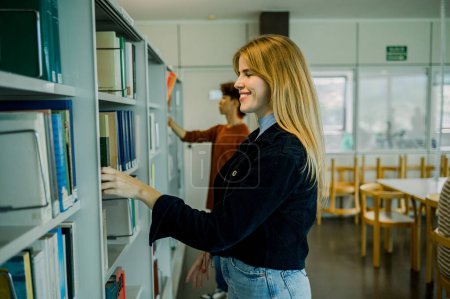 Téléchargez les photos : Vue latérale de la jeune étudiante heureuse en vêtements décontractés debout près de la bibliothèque dans la bibliothèque et la sélection de livres tout en passant du temps à la recherche pendant la journée - en image libre de droit
