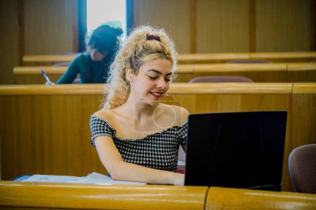 Téléchargez les photos : Jeune étudiante blonde positive en vêtements décontractés assise au bureau à côté d'un ordinateur portable et regardant l'écran tout en se penchant latéralement et en naviguant pour obtenir des informations en classe - en image libre de droit