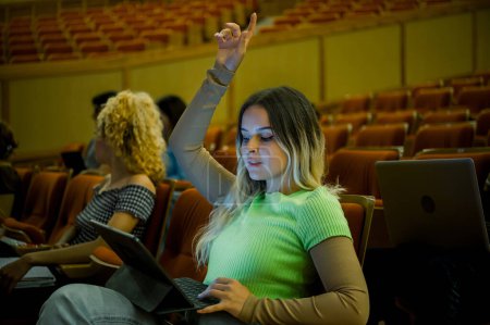 Foto de Vista lateral de una joven estudiante con ropa casual sentada con computadora portátil en el auditorio y levantando la mano mientras aprende - Imagen libre de derechos
