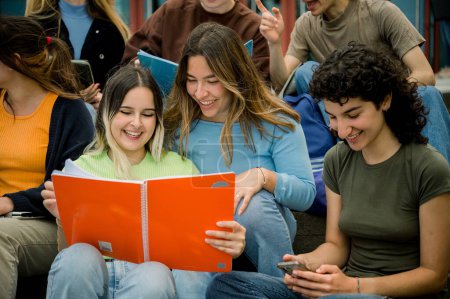 Téléchargez les photos : Groupe d'amis heureux de collège multiethnique avec des manuels et un smartphone se préparant pour le test et souriant tout en apprenant ensemble sur des bancs - en image libre de droit